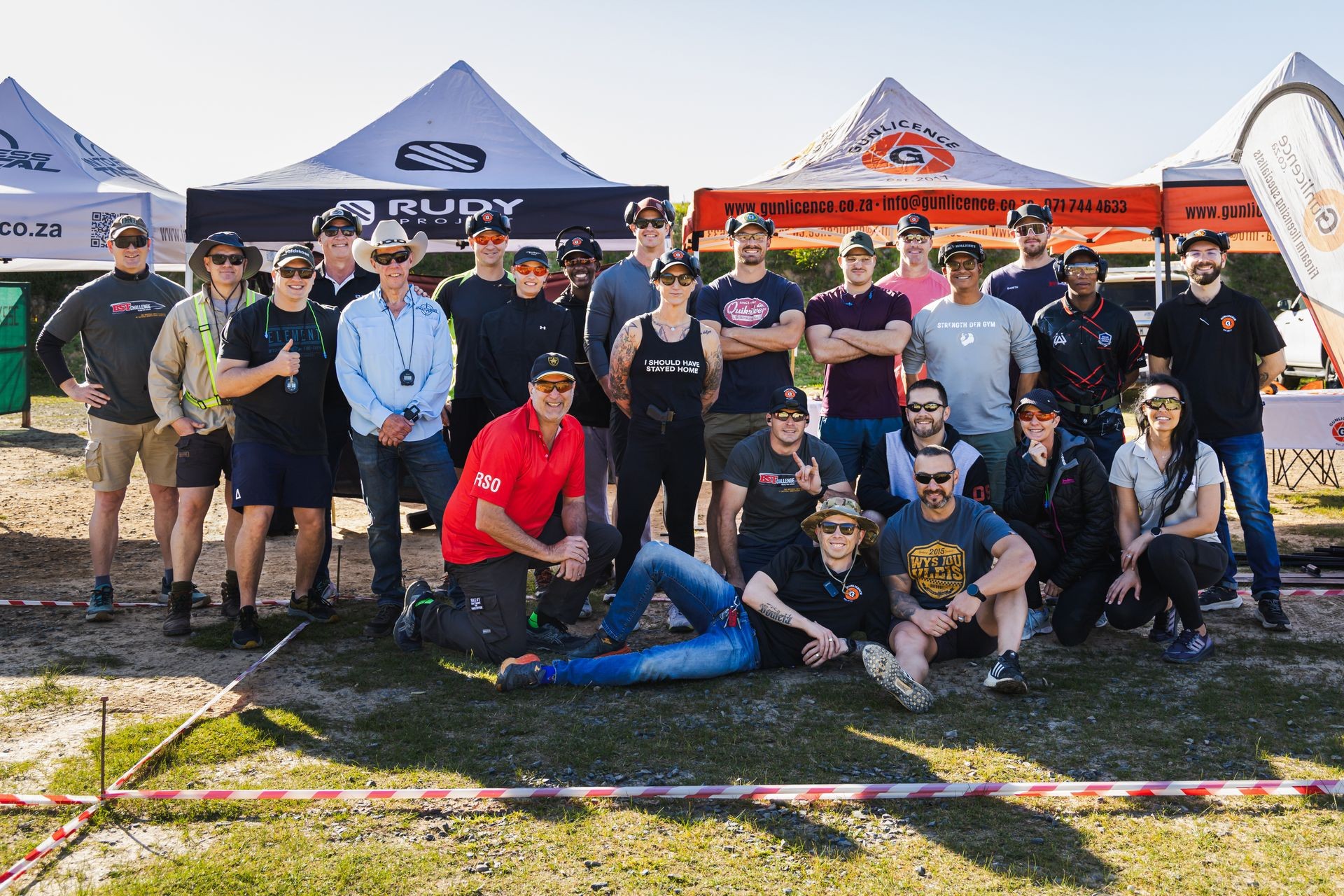 Group of people posing together outdoors in front of branded tents, some wearing sunglasses and casual attire.