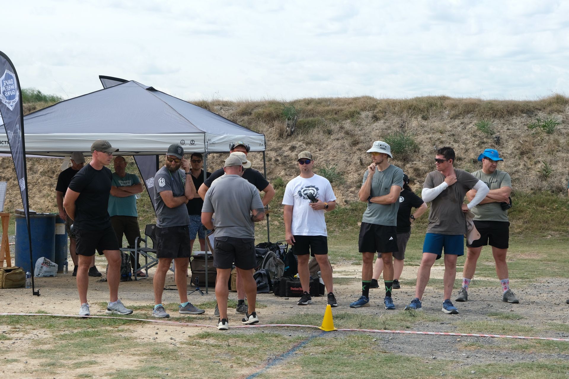 Group of men standing under a tent, listening intently outside on a grassy area.