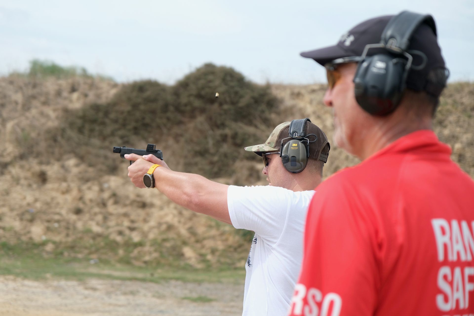 Person with protective gear shooting a handgun at a range under supervision.