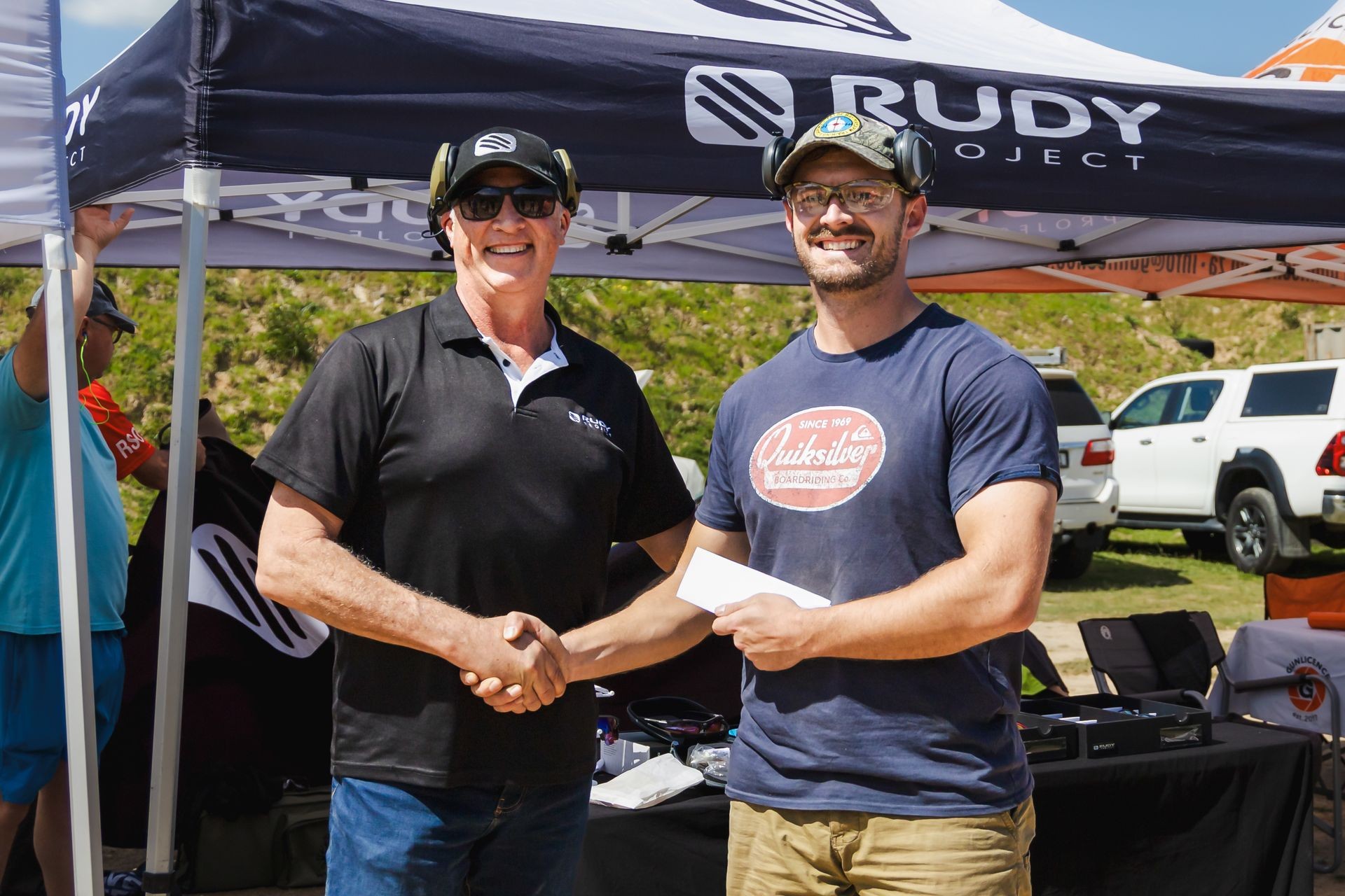 Two men shaking hands under a Rudy Project tent at an outdoor event, one holding an envelope.