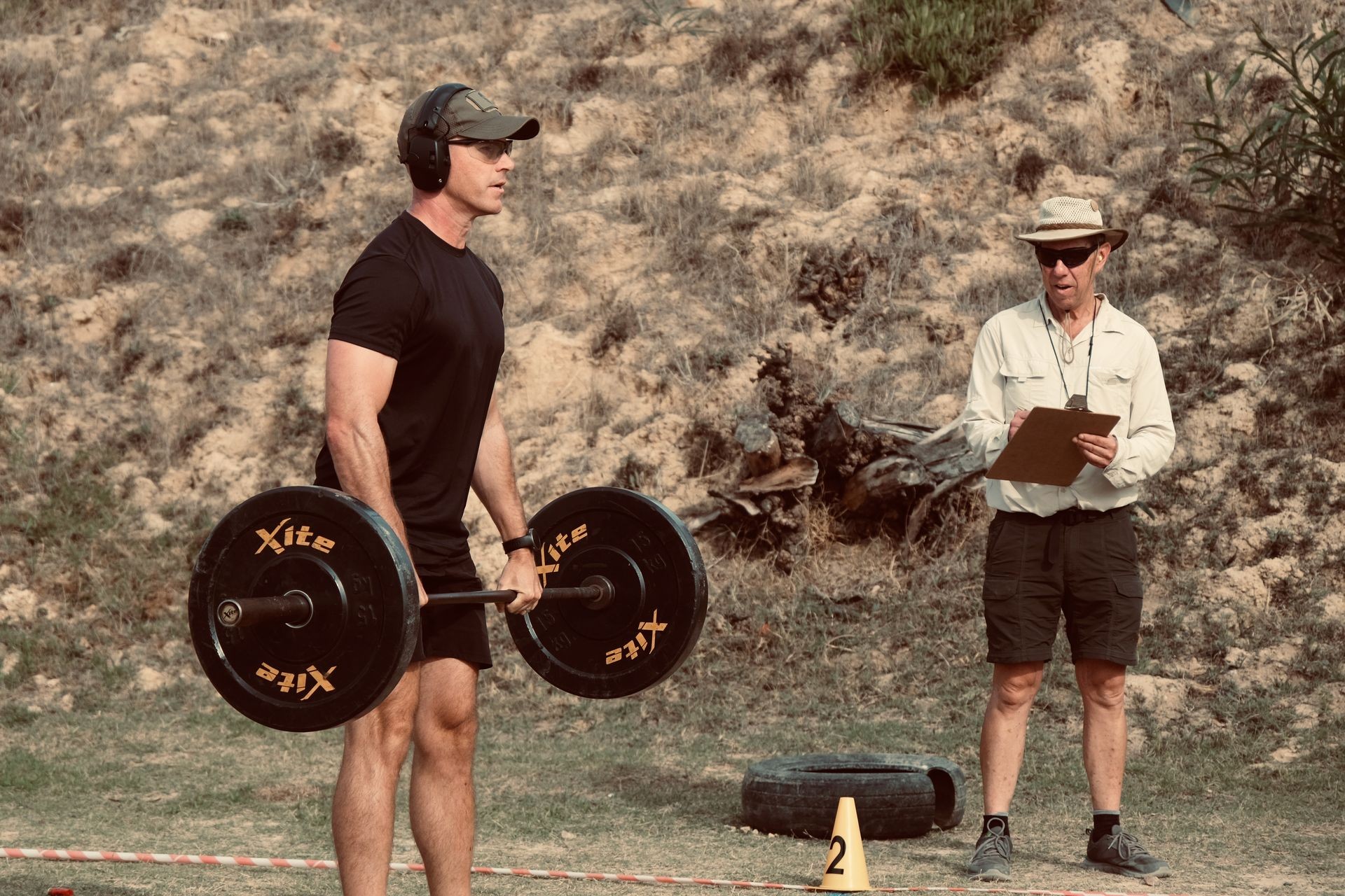 Man deadlifting a barbell outdoors under supervision of an instructor holding a clipboard.