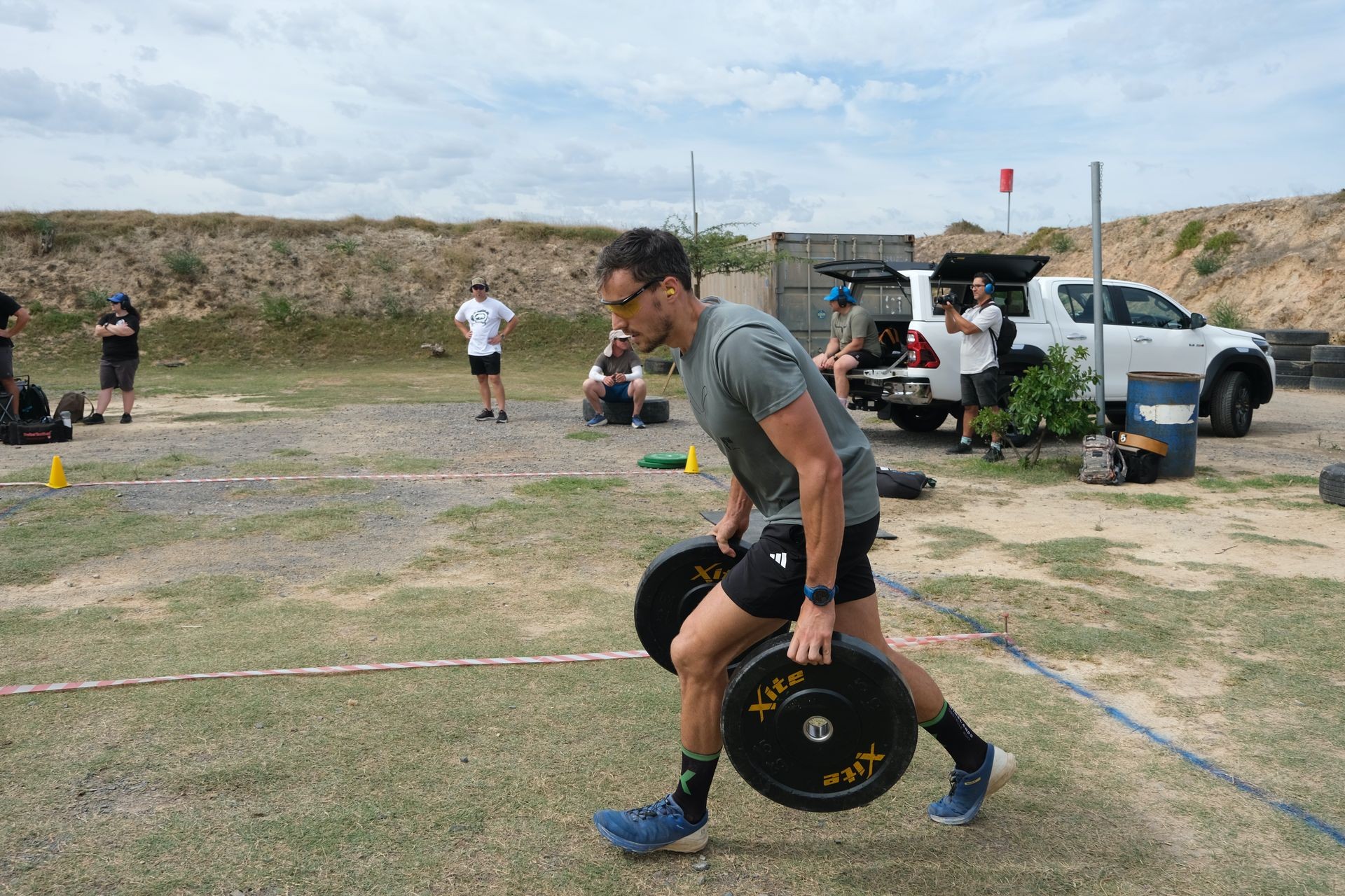 Man carrying heavy weights in an outdoor competition, with spectators and a truck in the background.