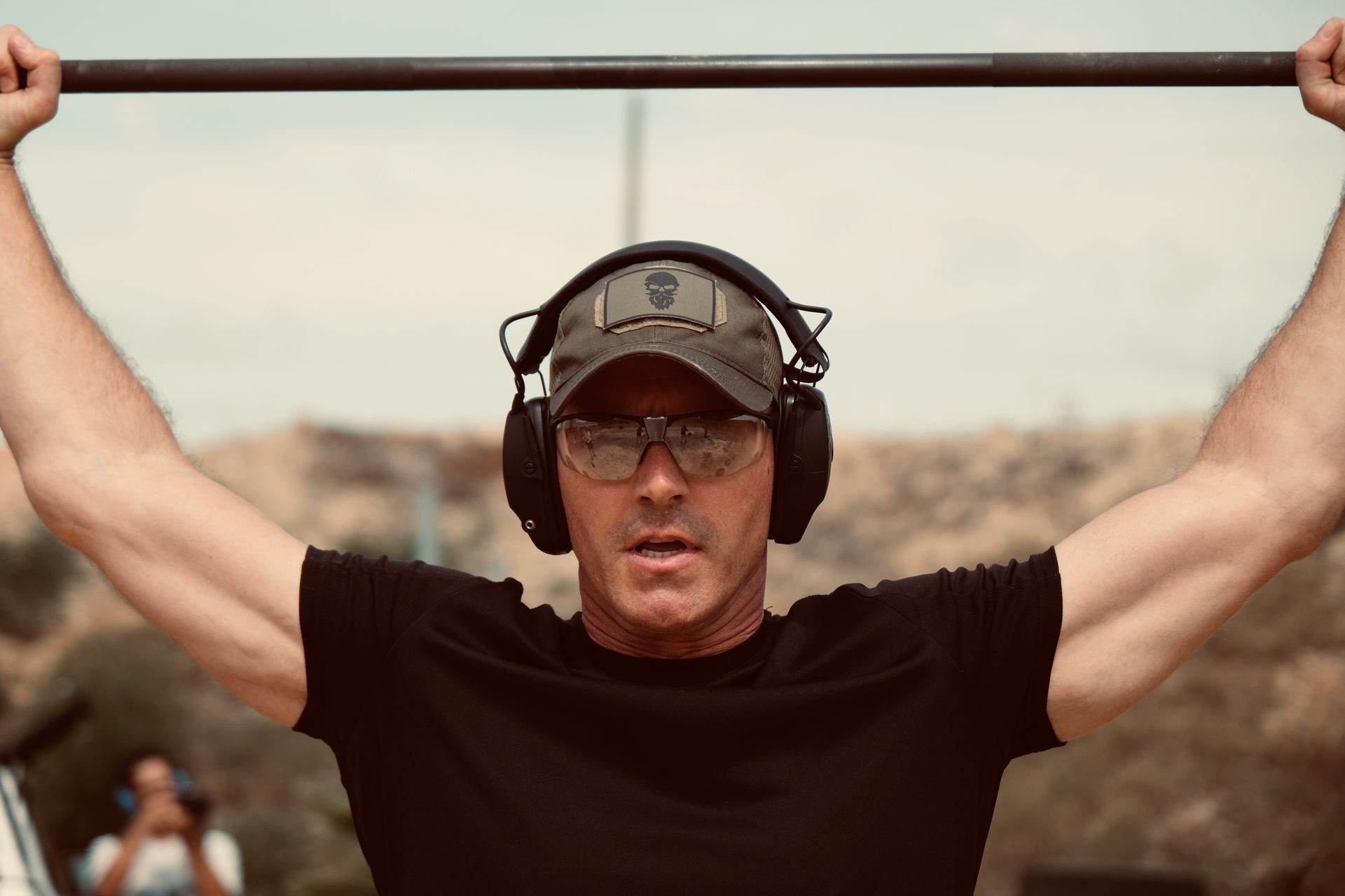 Person in black shirt and cap, wearing sunglasses and earmuffs, lifting a bar outdoors.