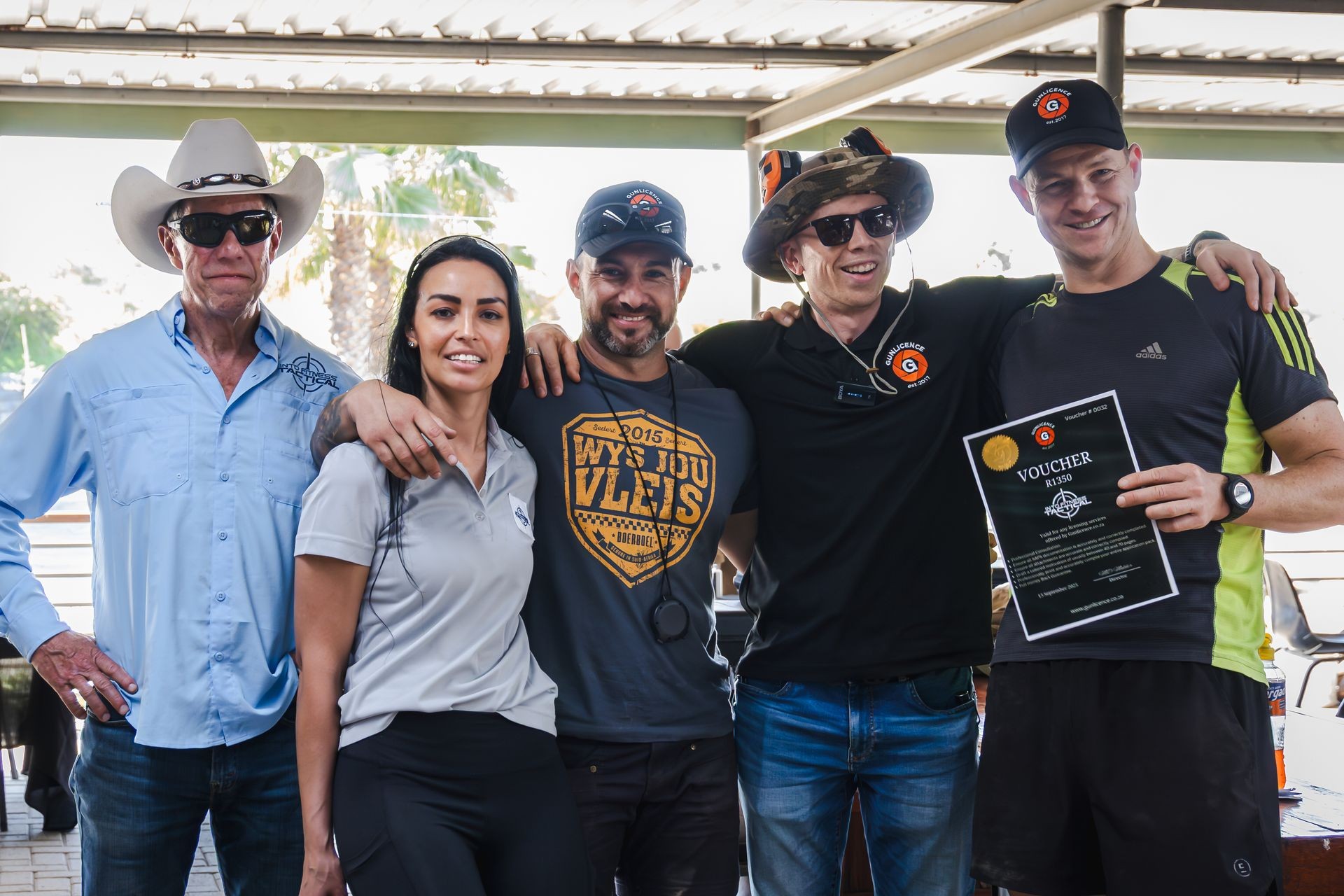Group of five people posing together, one holding a voucher, under a sheltered outdoor area.